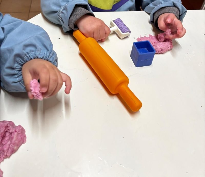 Manos de niñas y niños con guardapolvo celeste que juegan con masa rosa, un rodillo naranja y moldes sobre la mesa