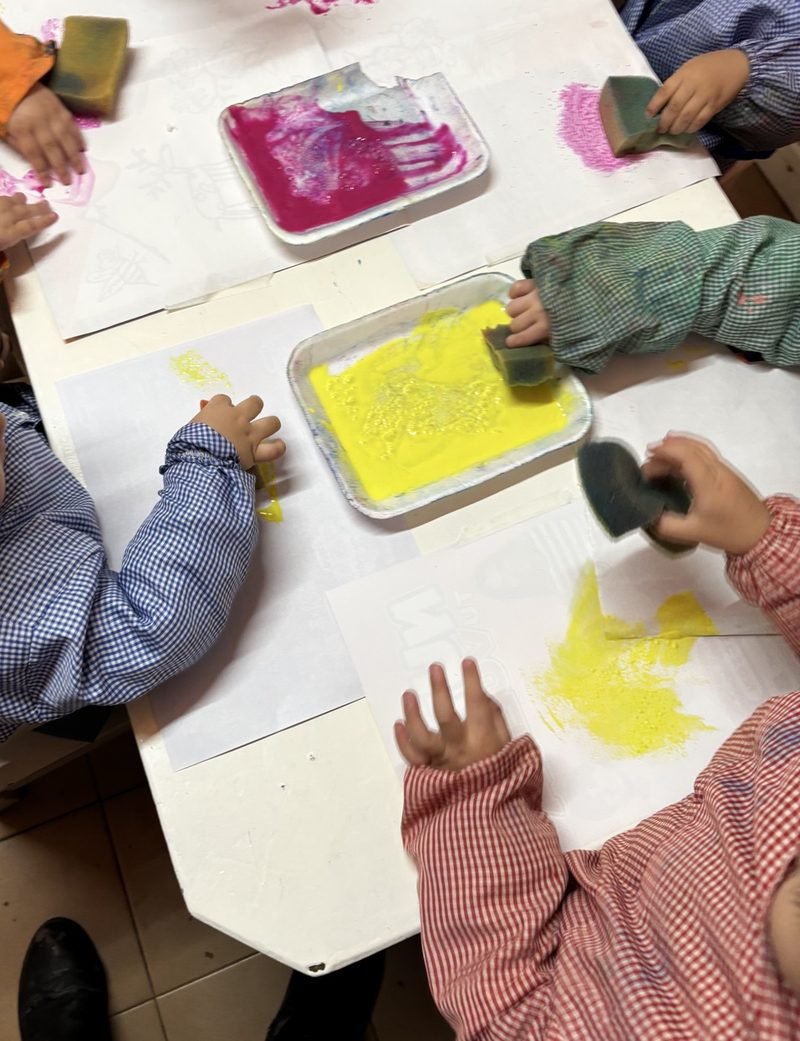 Manos de niñas y niños que estampan con esponjas pintura fucsia y amarilla sobre hojas blancas