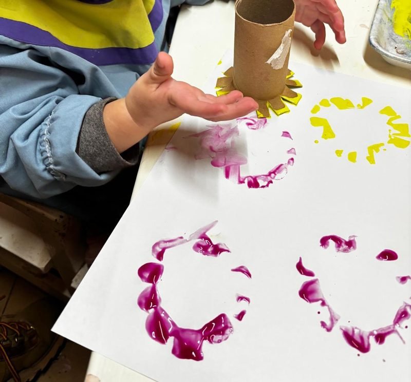 Mano de un niño que estampa círculos de pintura fucsia y amarilla con un rollo de cartón recortado